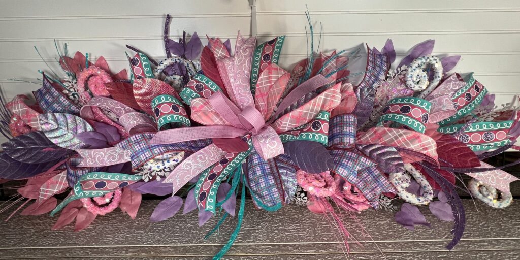 Horizontal floral swag made with the DecoExchange December 2025 wreath supply box, featuring layered pink, purple, teal, and patterned ribbon, glitter accents, faux leaves, and a centered bow for a winter or Easter display.