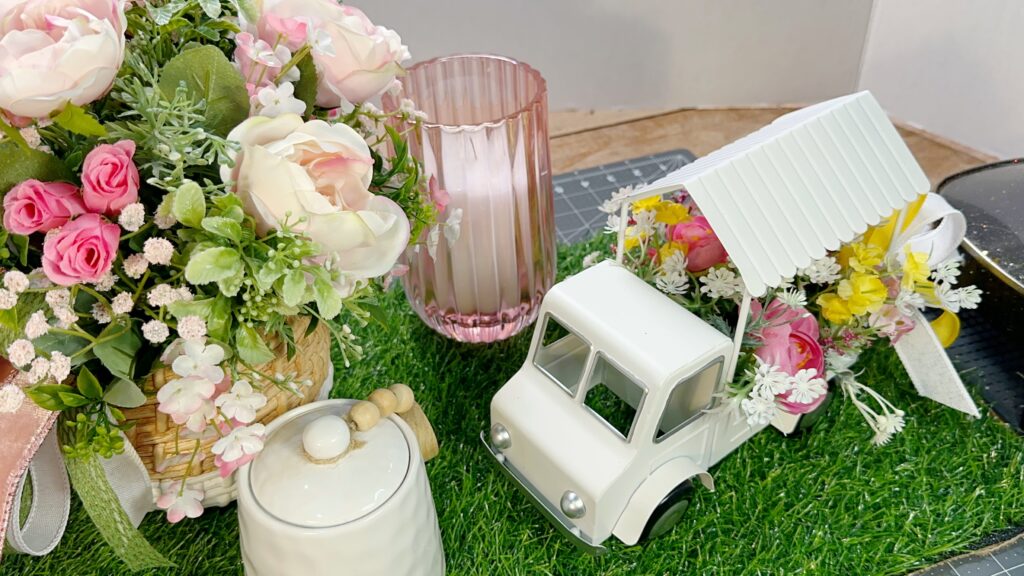 Close up of a pastel floral arrangement with pink and cream roses, greenery, a pink ribbed candle holder, a small white ceramic jar, and a white decorative truck filled with yellow and pink spring flowers displayed on faux grass.