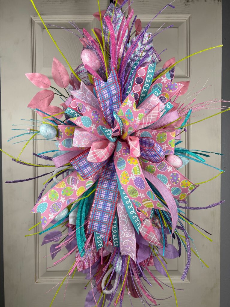 Vertical Easter-themed floral swag created with the DecoExchange December 2025 wreath supply box, showcasing pastel pink, teal, and purple ribbons, decorative Easter eggs, glitter sprays, and textured greenery on a door.