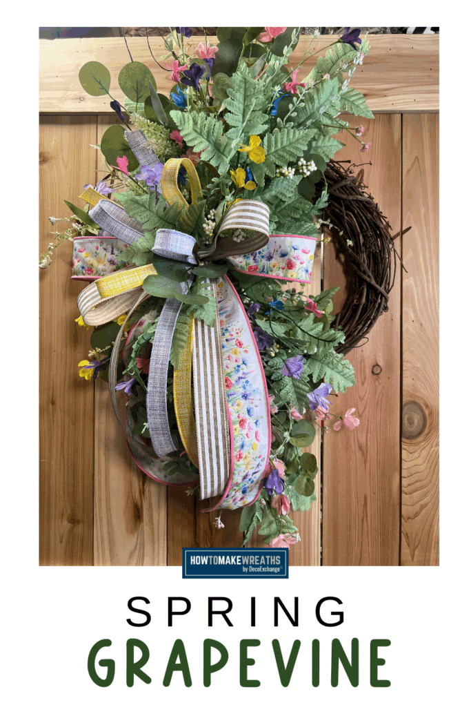 Close up of a spring grapevine wreath with pastel ribbon bow, mixed greenery, and colorful faux flowers on a wooden background.