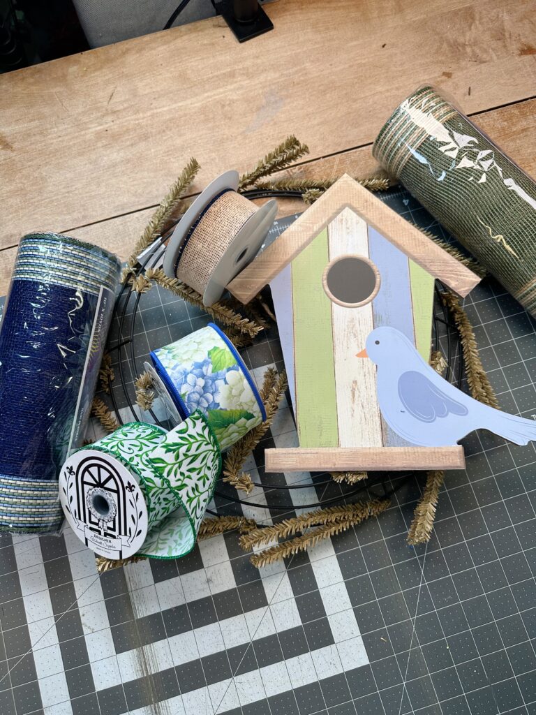 Supplies for making a spring birdhouse mesh wreath including deco mesh, ribbon rolls, wire wreath frame, and birdhouse sign on craft table.