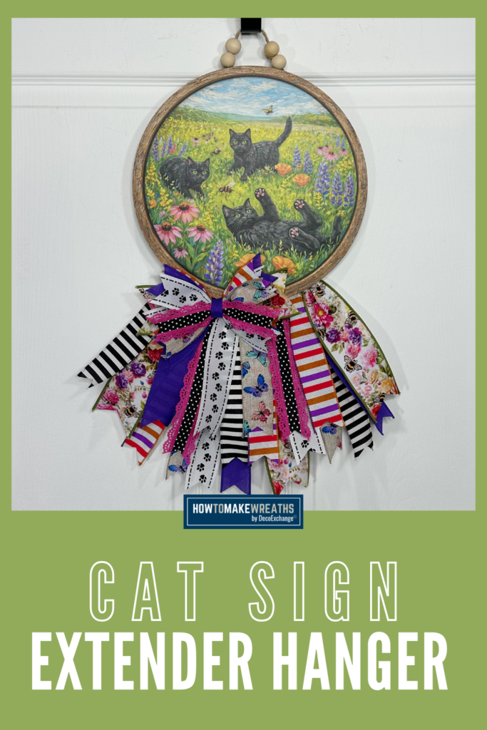 A round wood sign featuring black cats in a spring flower field, decorated with colorful layered ribbon loops in floral, stripe, polka dot, paw print, and butterfly patterns, hanging on a white door with wooden bead accents.