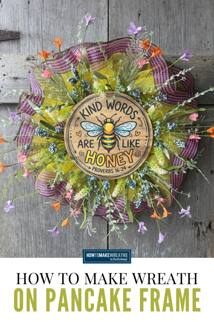 Small pancake wreath made with yellow mesh, pink and black striped mesh, wildflower florals, and a round wood bee sign that says Kind Words Are Like Honey hanging on a rustic wood background.