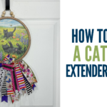 Tutorial thumbnail showing a round wood sign with black cats in a spring meadow, decorated with colorful layered ribbon tails and wooden beads, hung on a white door beside bold text reading How to Make a Cat Sign Extender Hanger.