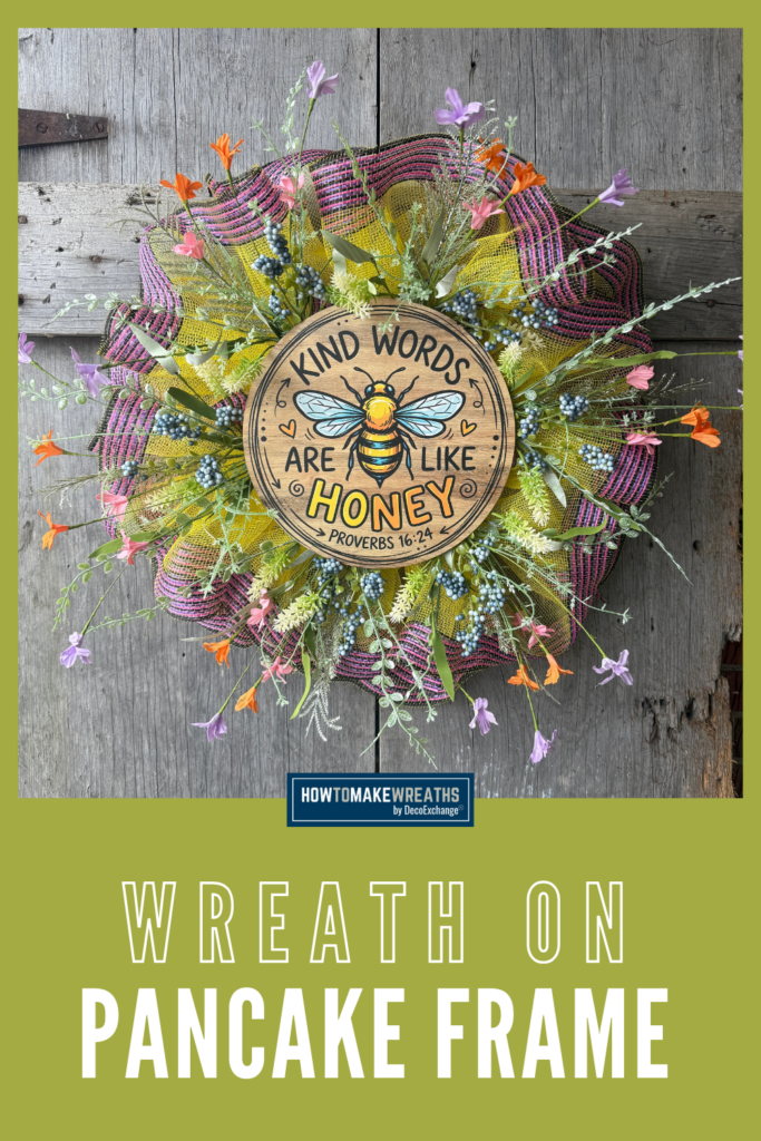 Small pancake wreath made with yellow mesh, pink and black striped mesh, wildflower florals, and a round wood bee sign that says Kind Words Are Like Honey hanging on a rustic wood background.