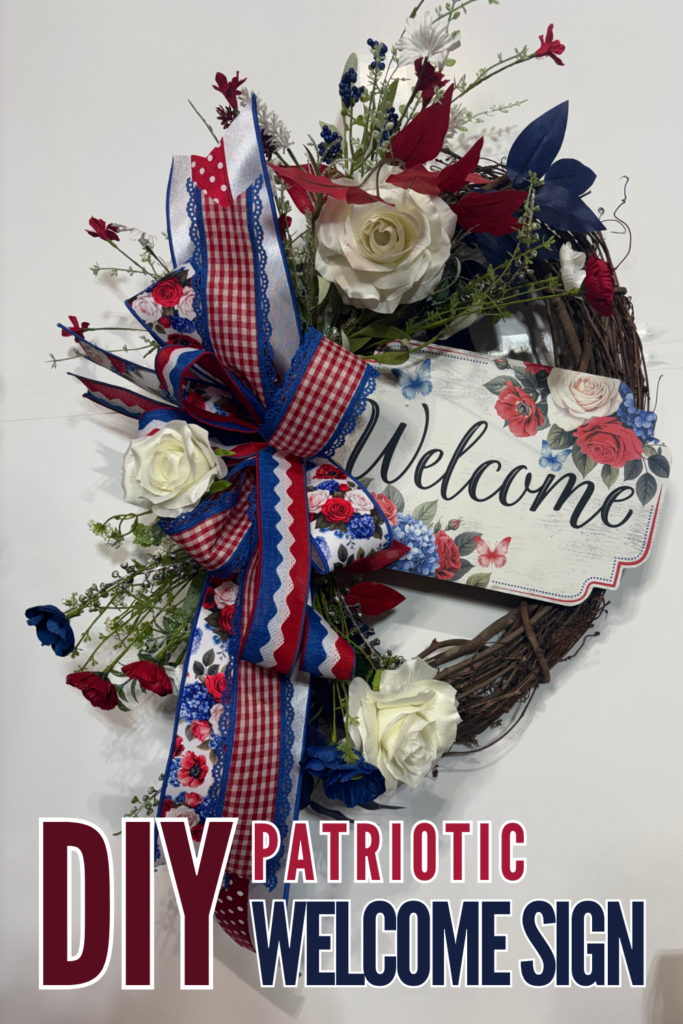 A traditional patriotic grapevine wreath with cream roses, red and navy blue silk florals, greenery, and a layered red, white, and blue gingham and floral ribbon bow, with a floral welcome sign centered on the wreath, photographed against a white background.