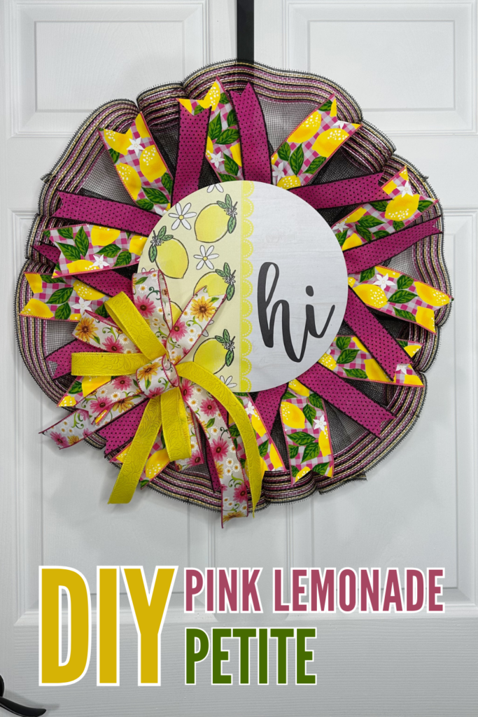 A round pancake petite wreath made with black deco mesh and layered pink, yellow, and lemon-print ribbon tails, with a yellow floral bow and a round hi lemon sign centered on the wreath, hung on a white door.