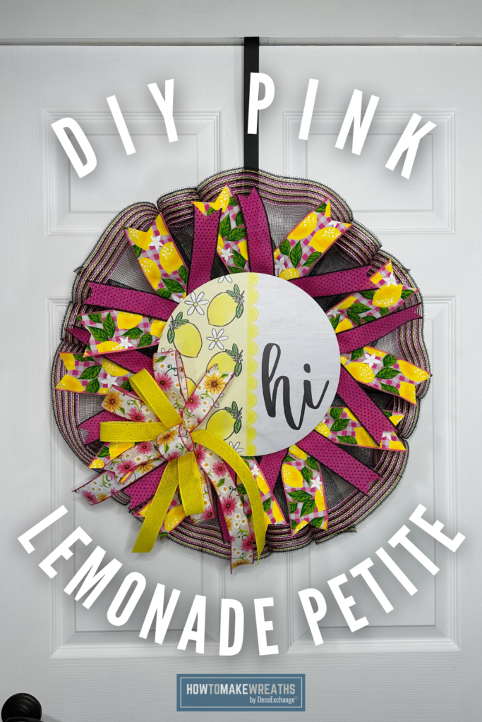 A pink lemonade pancake petite wreath with black deco mesh, alternating hot pink and lemon-print ribbon loops, a yellow floral bow, and a round lemon hi sign in the center, hung on a white paneled door with a black over-the-door hook.