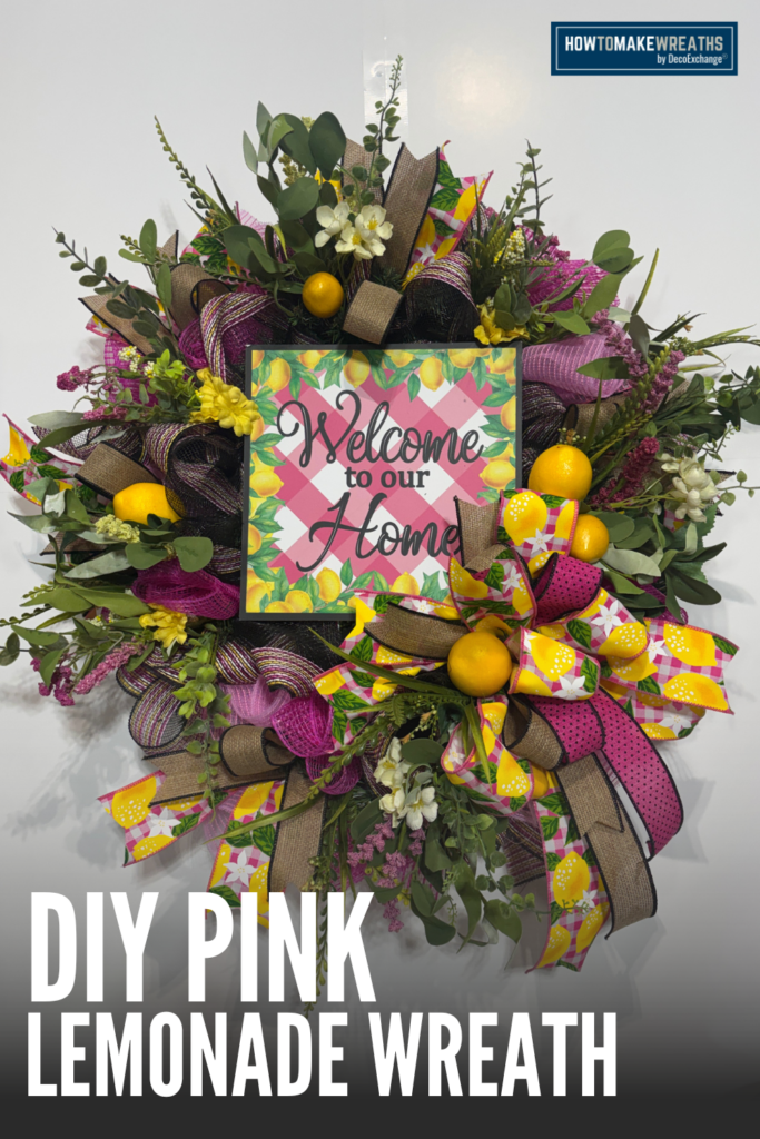 A full round evergreen wreath covered in hot pink and black deco mesh with layered loops of lemon print gingham, tan burlap, and pink polka dot ribbon, styled with yellow lemon ball picks, white blooms, small yellow flowers, eucalyptus sprigs, and purple filler stems, with a square Welcome to our Home sign at the center featuring a pink gingham and lemon border, hung against a white wall.
