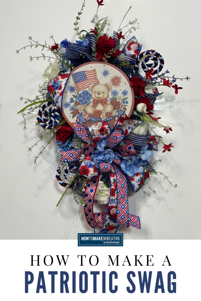 A patriotic swag made with white deco mesh, red and blue silk florals, pinecone picks, and a round wooden bear sign featuring an American flag, finished with a layered red, white, and blue star and floral ribbon bow with long tails, displayed on a white wall.