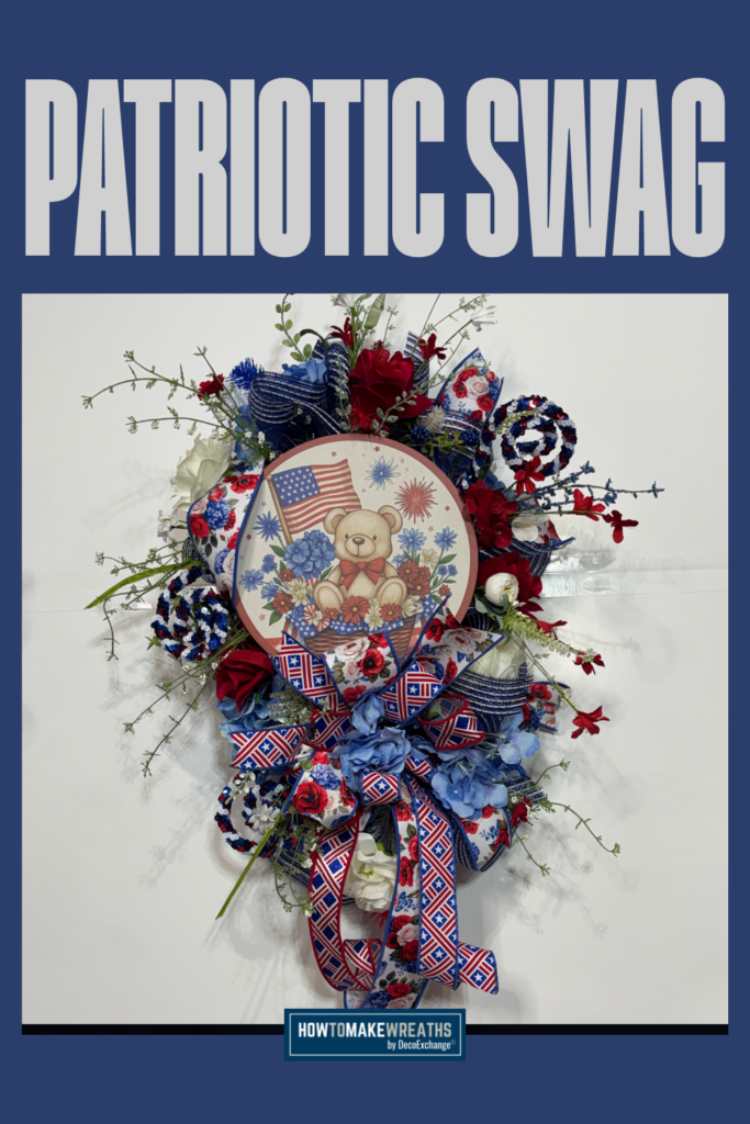 A patriotic swag with white deco mesh, red roses, blue hydrangeas, red and blue floral picks, pinecone accents, and a round bear and American flag wood sign, finished with a star-print and floral ribbon bow, shown on a white wall with a navy blue border.