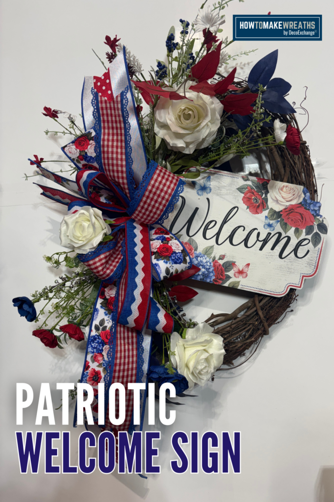 A patriotic grapevine wreath decorated with cream roses, red and blue silk florals, greenery, and a layered red, white, and blue gingham and floral ribbon bow, with a welcome sign featuring roses and butterflies nestled in the center.