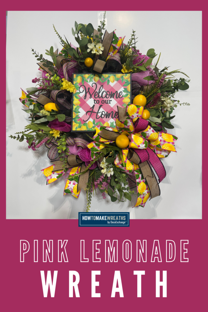 A large round evergreen wreath base covered in hot pink and black deco mesh with looped lemon gingham, pink polka dot, and tan burlap ribbon, accented with realistic yellow lemon ball picks, small white florals, yellow blooms, eucalyptus, and purple filler stems, centered with a square Welcome to our Home metal sign framed in a pink gingham and lemon print border, displayed against a white wall.