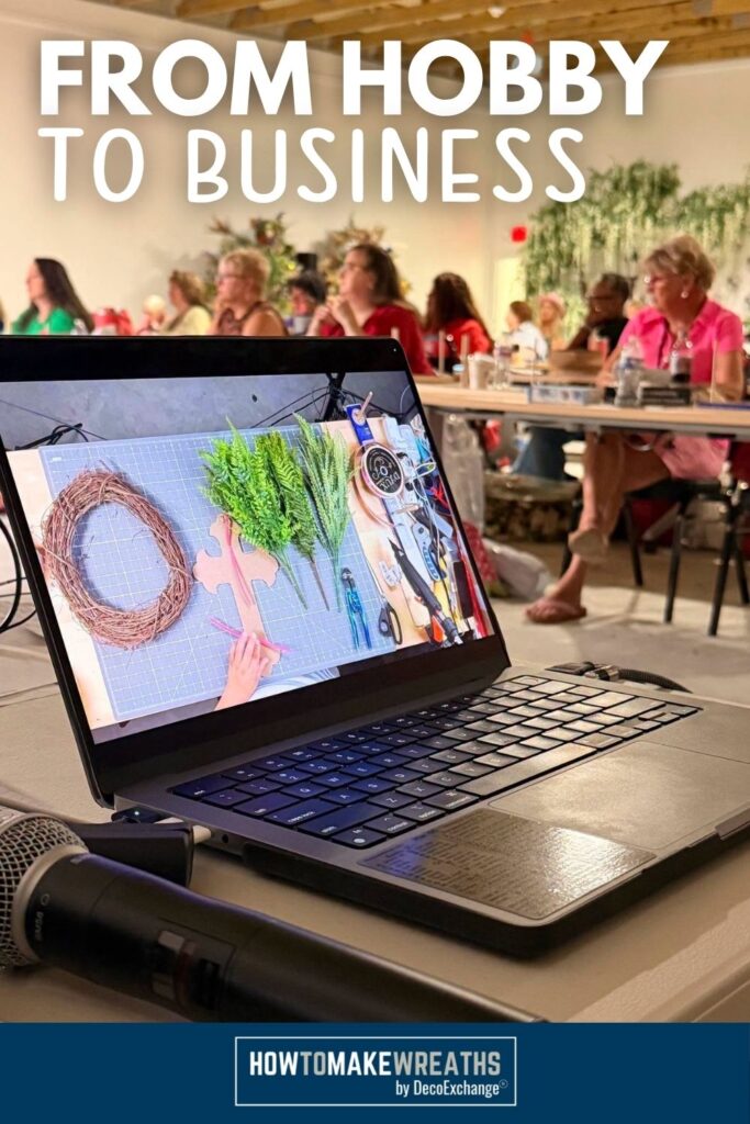 A laptop on a table displays a wreath-making tutorial video showing a grapevine wreath base, faux greenery stems, and craft tools laid out on a cutting mat, with a room of women working on wreaths in the background at a DecoExchange in-person workshop. Bold white text overlay reads "From Hobby to Business." Navy footer bar with the HowToMakeWreaths by DecoExchange logo.