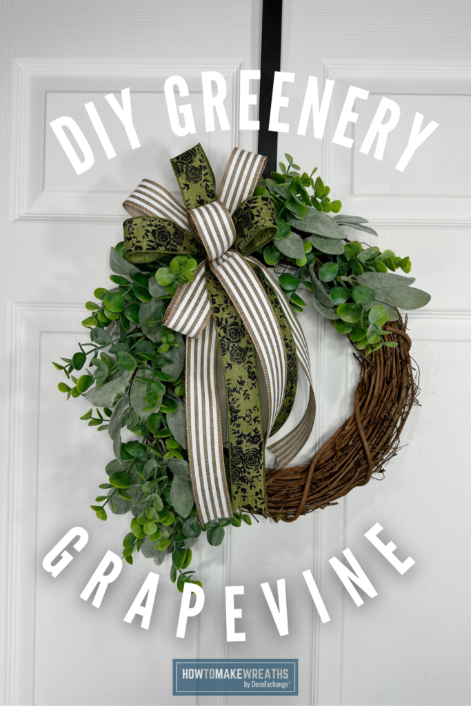 A round grapevine wreath covered in mixed green eucalyptus and lambs ear leaves, topped with a bow made of green floral print ribbon and brown and white striped ribbon, hanging on a white paneled door.