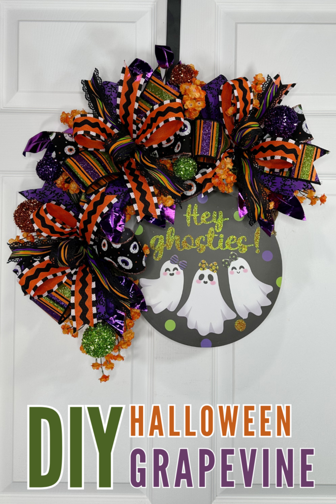 A full Halloween grapevine wreath built with a cascading layered bow in eyeball print, black and orange stripe, purple metallic, and multicolor wave ribbon, styled with orange baby's breath filler, metallic purple ficus, and glitter ball picks in green, orange, and rust, centered with a round dark sign reading Hey Ghosties featuring three smiling ghost characters, displayed on a white paneled door with a black ribbon hanger.