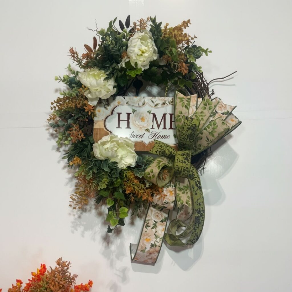 A grapevine wreath covered in layered deep green and rust-toned mixed greenery, eucalyptus, and fern stems, accented with three large white peony blooms, centered with a Home Sweet Home sign.