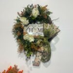 A grapevine wreath covered in layered deep green and rust-toned mixed greenery, eucalyptus, and fern stems, accented with three large white peony blooms, centered with a Home Sweet Home sign.