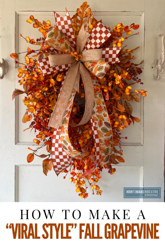 A fall grapevine swag with a teardrop shape, decorated with a large bow of rust and white checkered ribbon, burlap ribbon, and leaf print ribbon, surrounded by orange and yellow florals and berry picks, hanging on an antique white door.