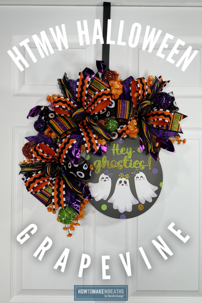 A Halloween grapevine wreath with a large layered bow in eyeball print, orange and black stripe, purple metallic, and wave ribbon, accented with orange baby's breath clusters and green, orange, and rust glitter ball picks, centered with a round dark sign reading Hey Ghosties with three cute ghost characters, hung from a black ribbon on a white paneled door.
