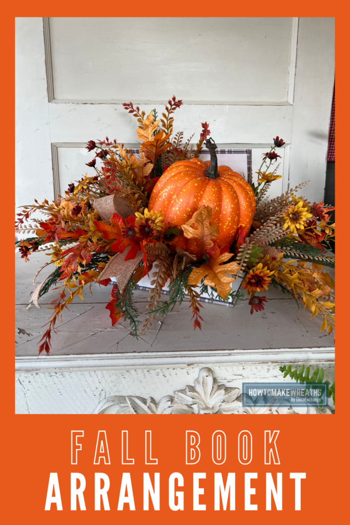 A fall centerpiece arrangement built on a faux book base with a speckled orange pumpkin, oak leaves, fern, fall daisies, pine picks, and a burlap ribbon bow, displayed on a distressed white mantel.